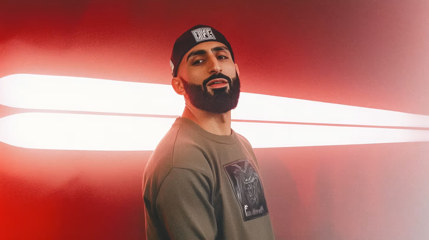 MMA fighter Belal Muhammad poses confidently with dark hat and olive-green shirt amid a dramatic red gradient background.