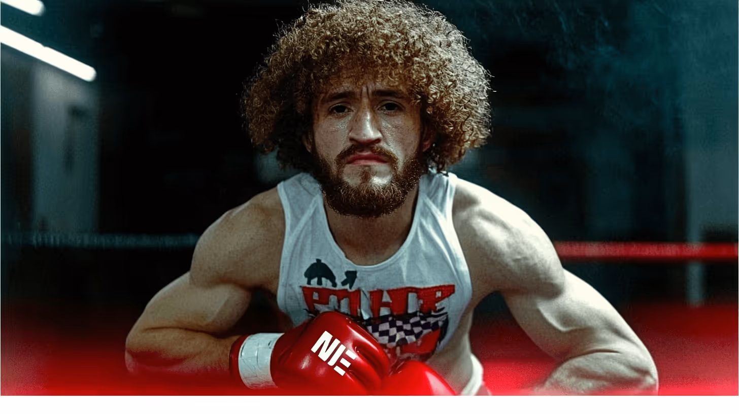 MMA fighter Merab Dvalishvili strikes a determined pose in a gym, wearing a sleeveless white shirt and bold red gloves.