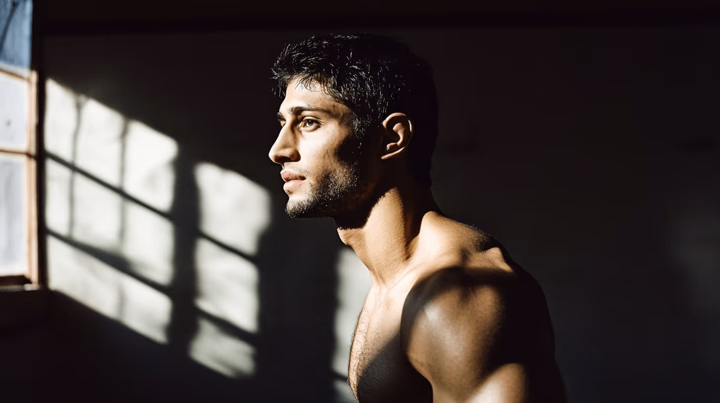 Allan Nascimento MMA fighter in side profile, bare-chested with muscular shoulders in a dimly lit room, exuding focus.