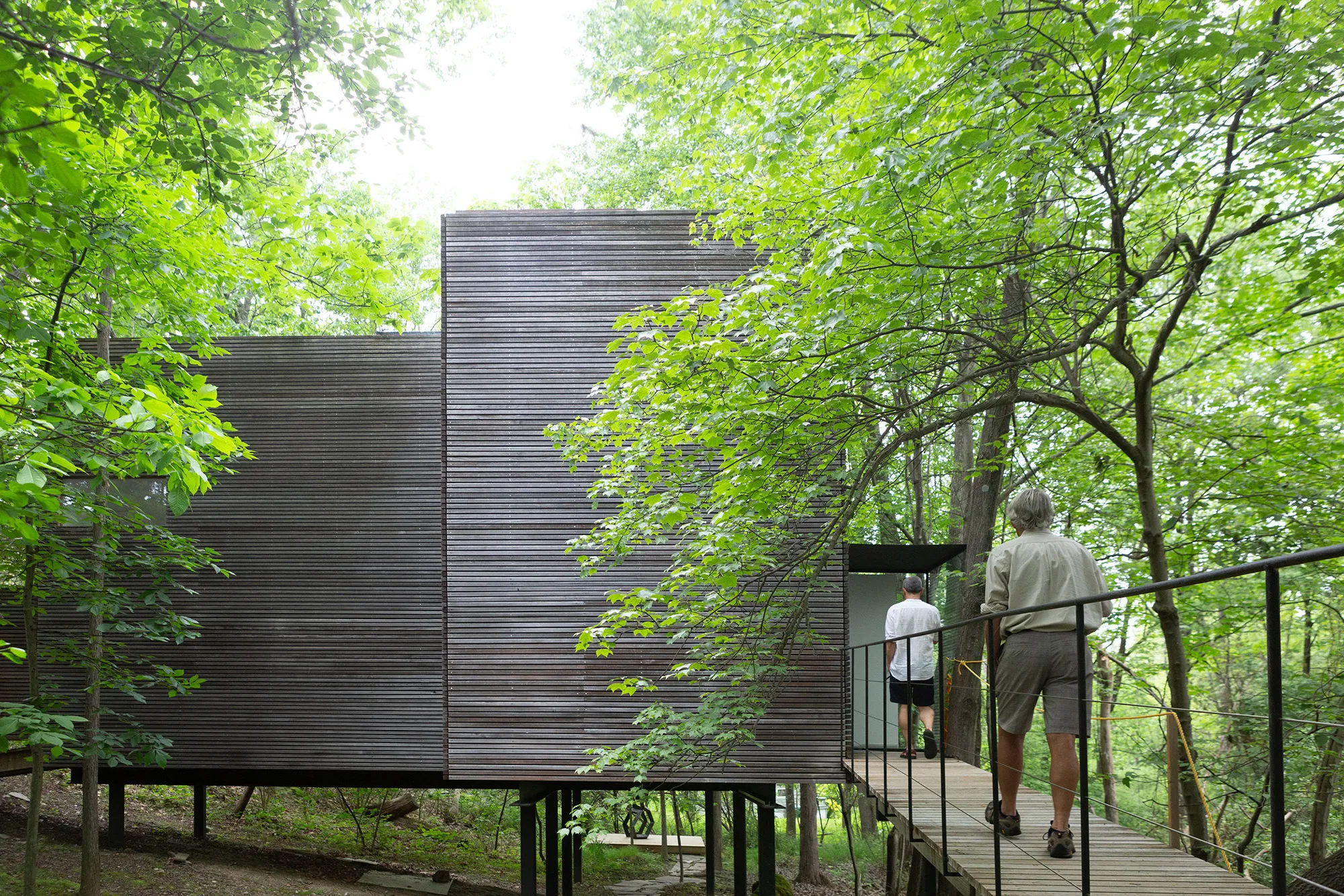 Two figures walking on a sky bridge towards an elevated one story building in the background.
