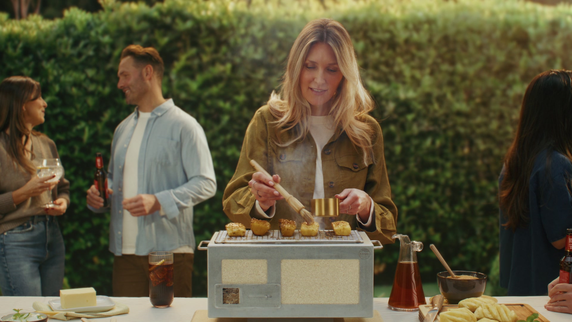 Smiling bearded man in cream shirt and mustard tee grilling on Kasai teppan in a green terrace-garden setting.