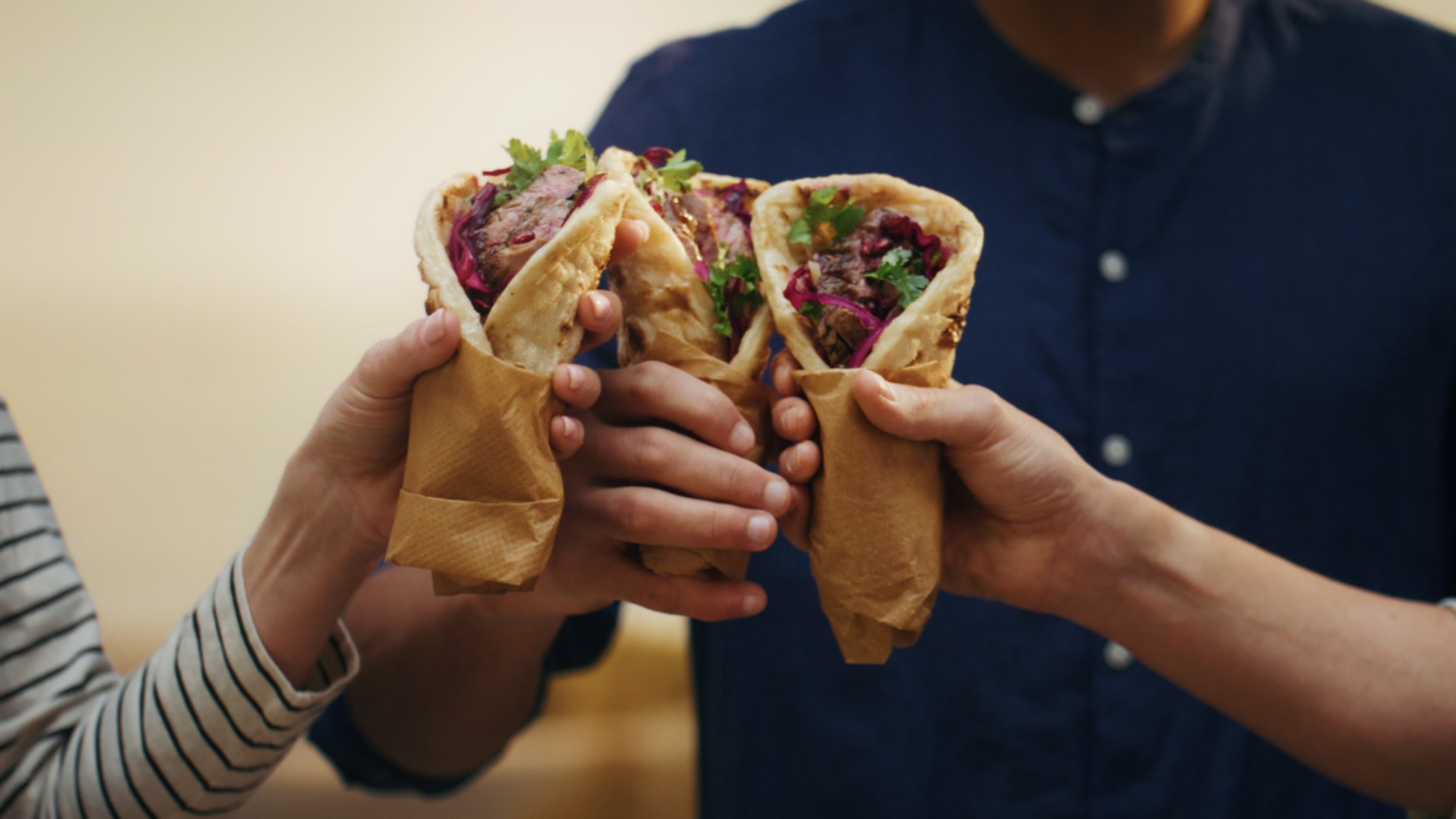 Three hands cheers with folded flatbreads stuffed with grilled lamb, pickled cabbage and herbs, in paper wraps.