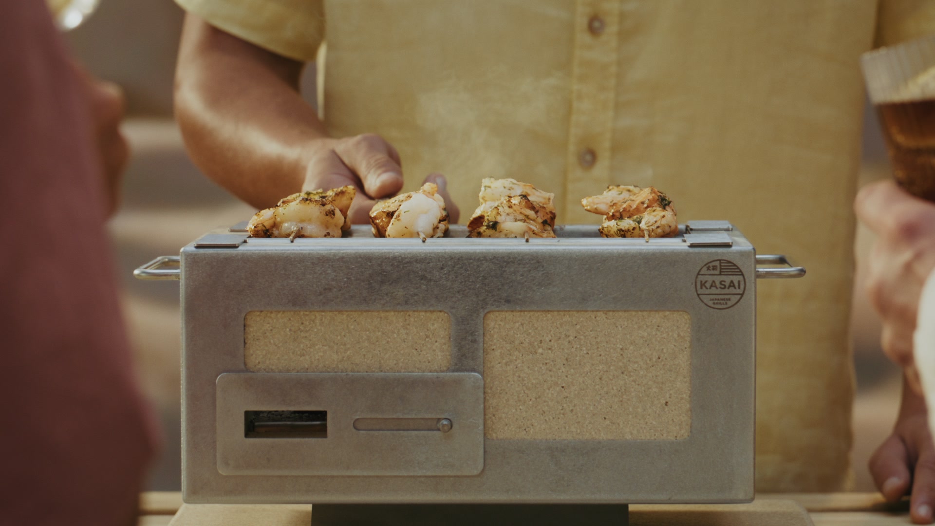 Blonde woman in olive jacket basting five seed-crusted bread rolls on a Kasai Konro grill with a gold bowl.