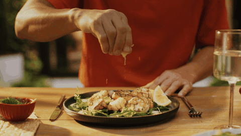 Hand squeezing a lemon half over grilled prawns; juice droplets frozen in mid-air; warm window bokeh.