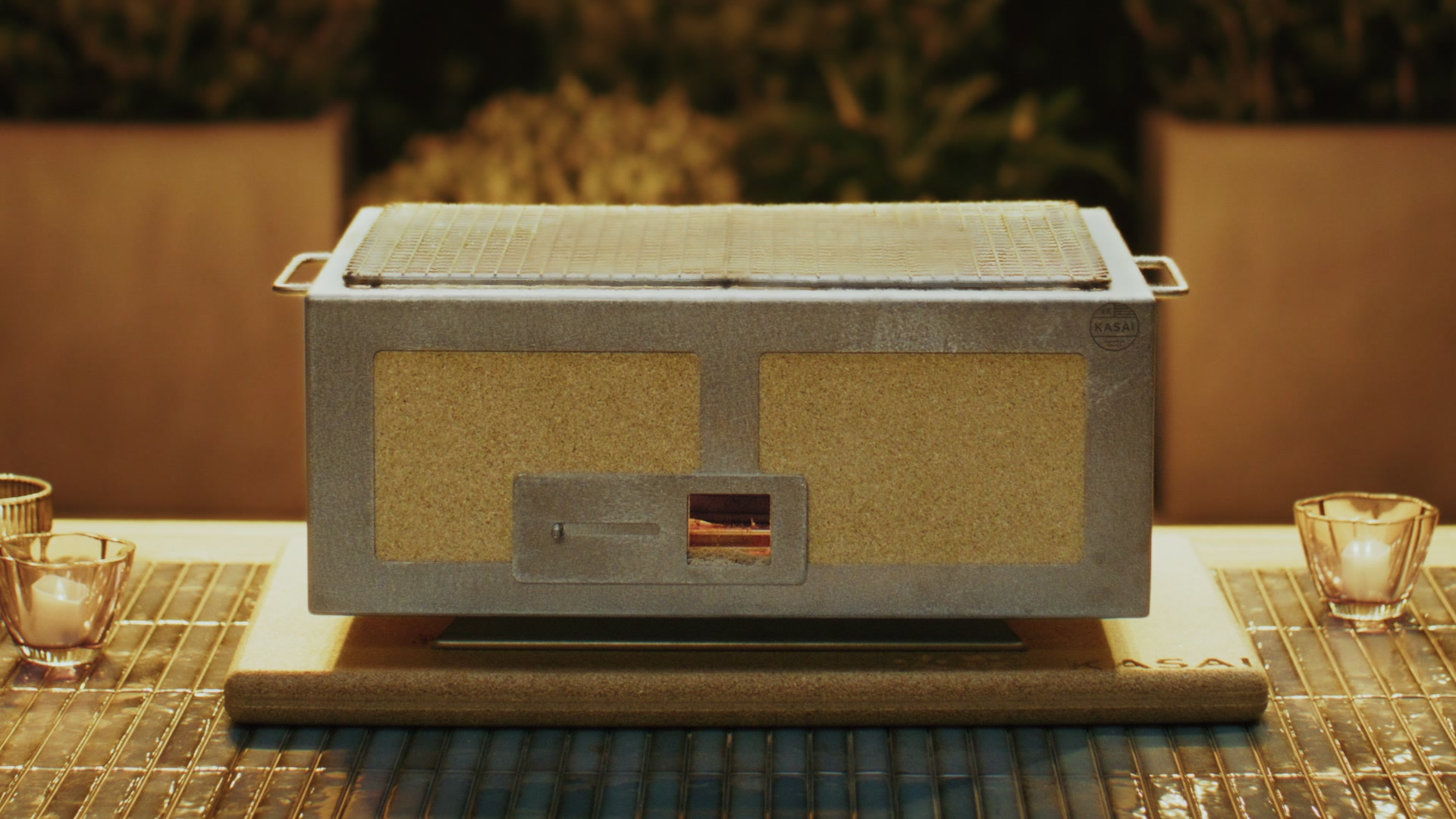 Kasai Konro grill with glowing red grate on a wooden table; lush green hedge and golden backlight behind.