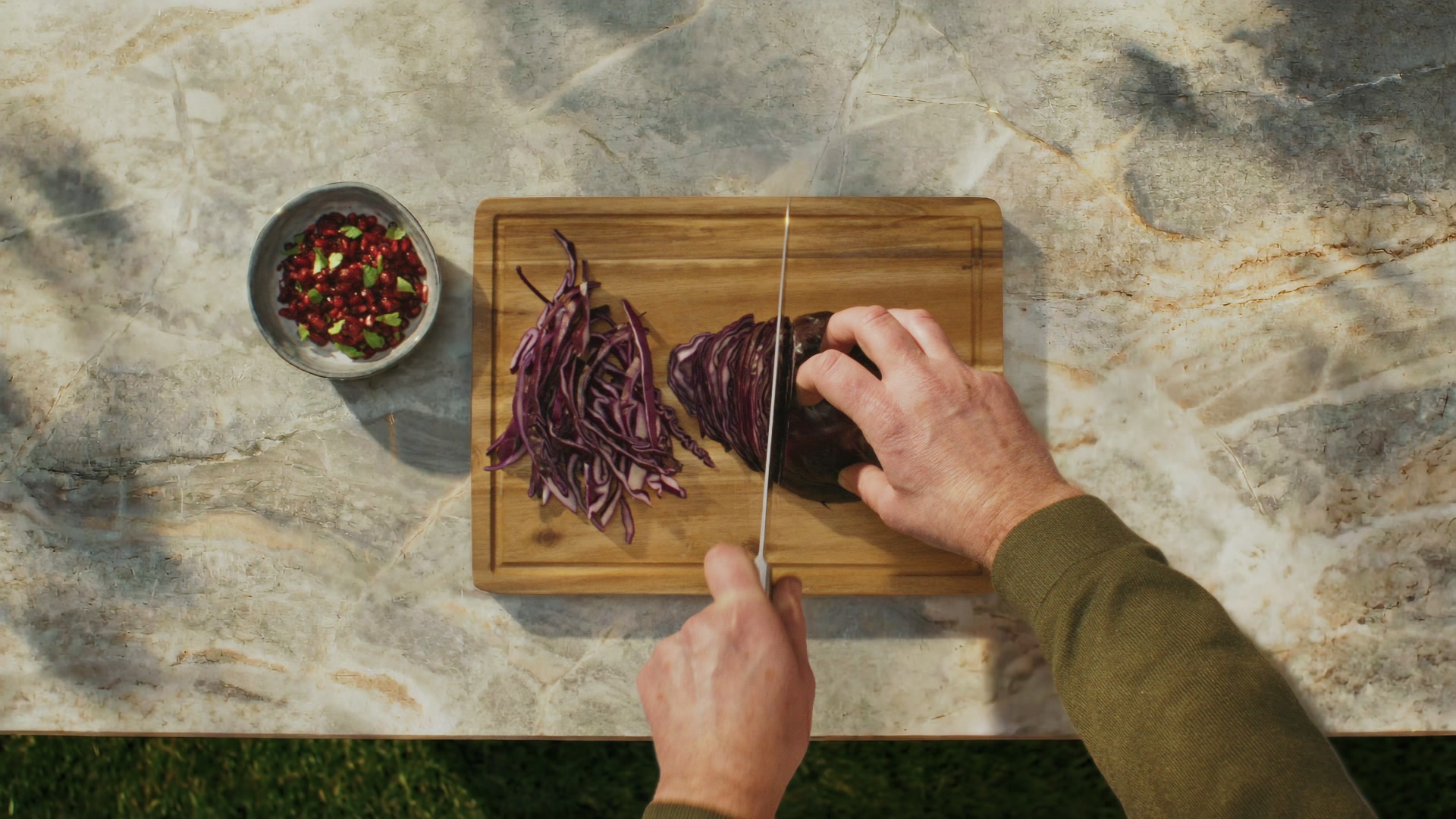 Overhead chopping board of fresh vegetables with a hand reaching for a red onion; warm stone surface.