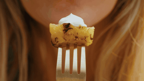 Macro close-up of grilled pineapple chunk with whipped cream on a gold fork, held to blurred lips.