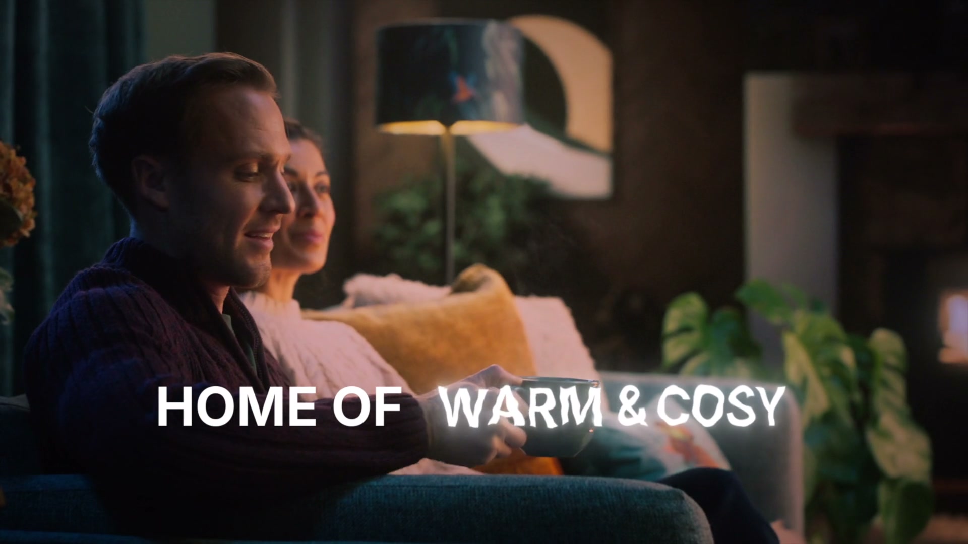 Couple cosied up on a sofa with a cuppa in a warmly lit cottage interior with HOME OF WARM AND COSY overlaid
