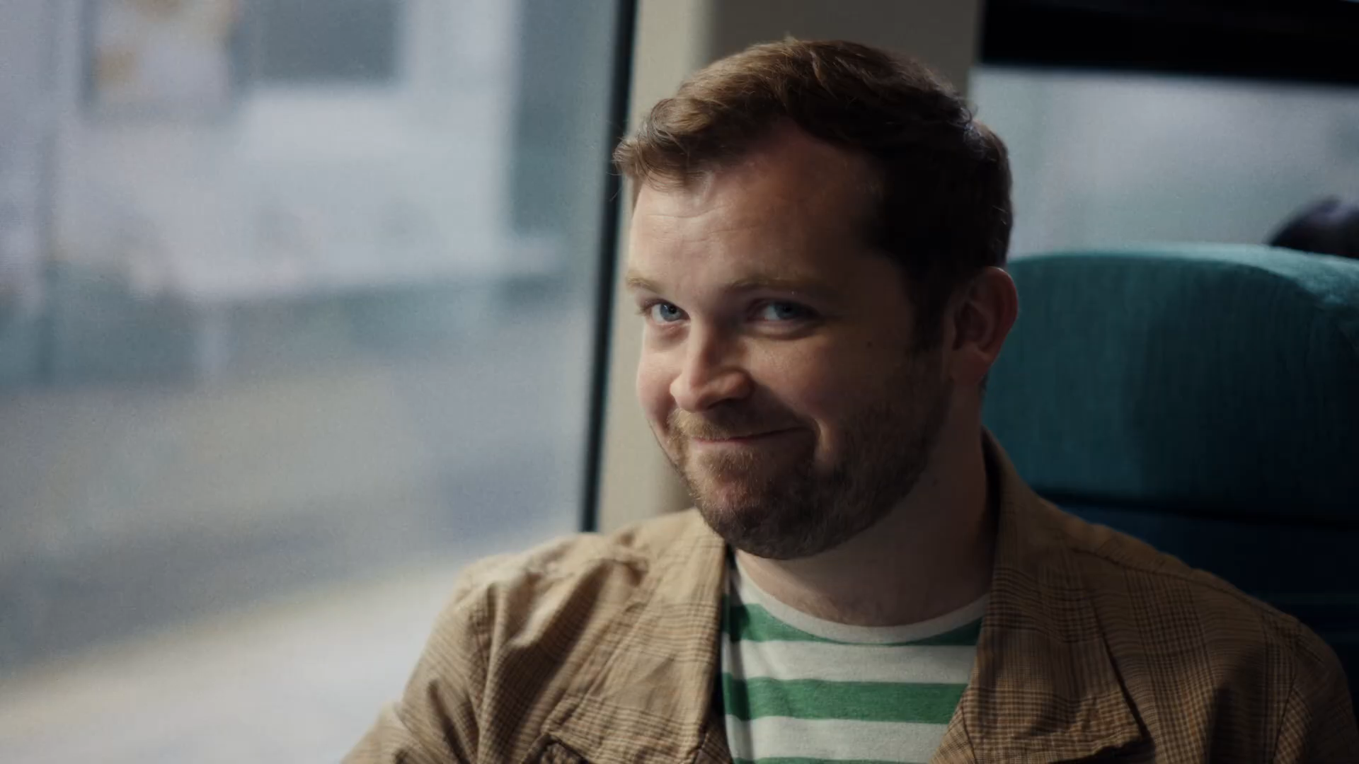 CU of Dad smirking conspiratorially toward Sacha off-camera, dimples showing — the 'Not today' playful look on the train