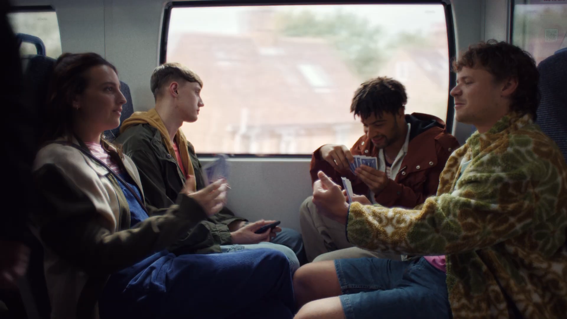 Friends playing cards on the Thameslink train — Lily, Luke, Billy and Callum at a table seat, soft countryside through the window