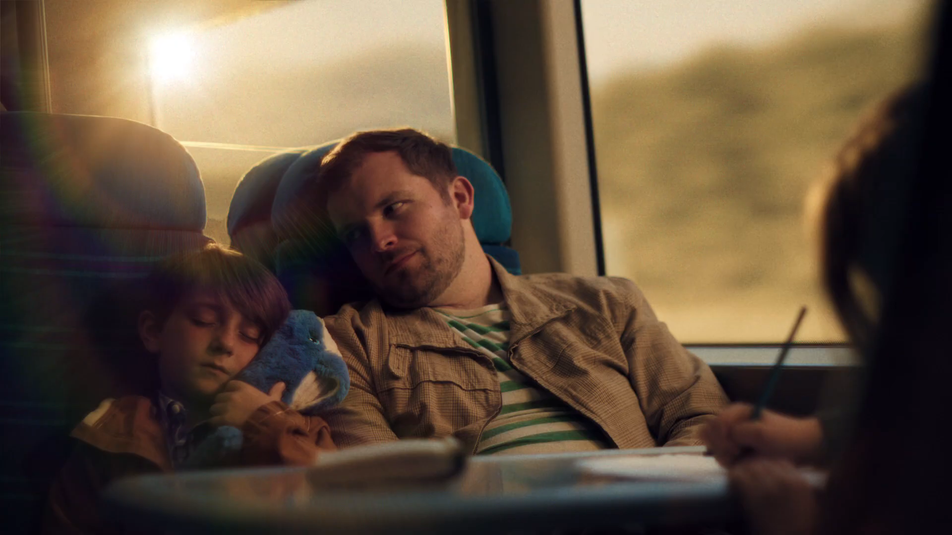 Cinematic train-home interior at golden hour — Lucas asleep cuddling a soft octopus toy souvenir, Dad beside him watching tenderly, Sacha drawing at the table seat