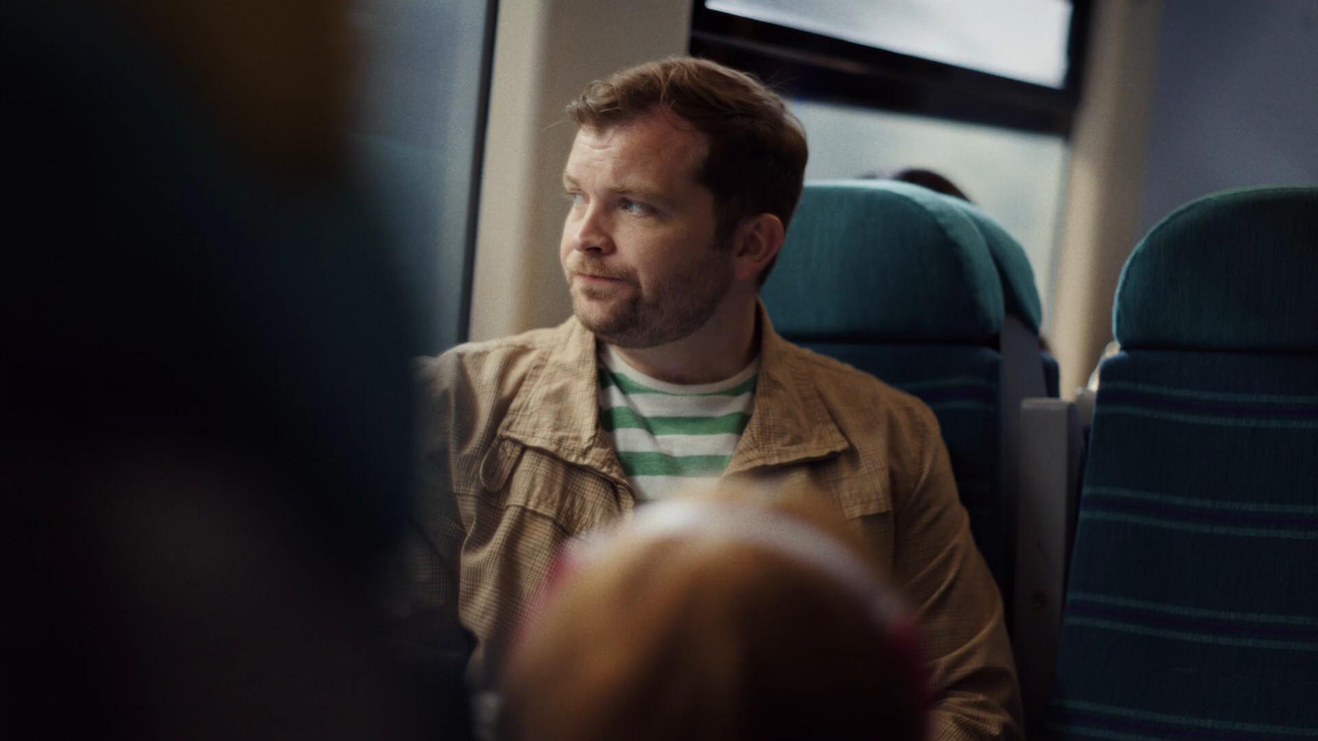 Cinematic MCU of Dad on the Southern train, profile-three-quarter, looking up the carriage past the camera, the panoramic train window glowing with daylight behind him