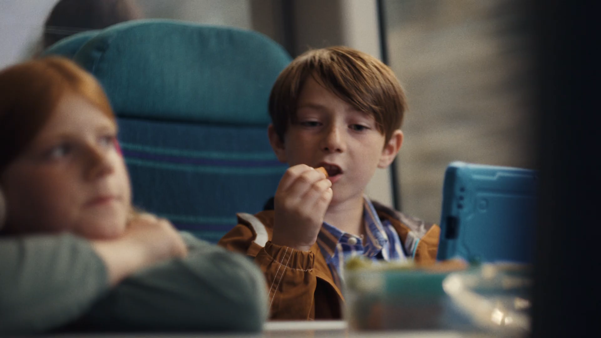 Two-shot of the kids at the train table seat — Sacha resting her chin on her crossed arms looking off-camera, Lucas right of frame eating snacks