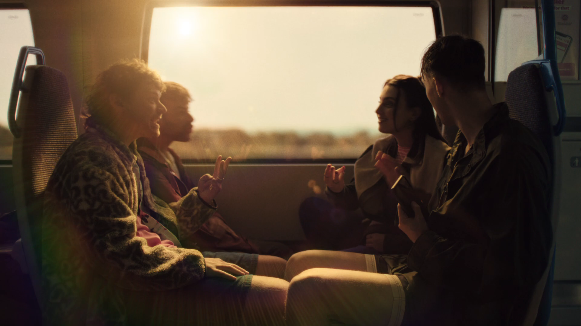 Golden hour resolution wide of the four friends sat across from each other on the Thameslink train home, sun blasting through the panoramic window