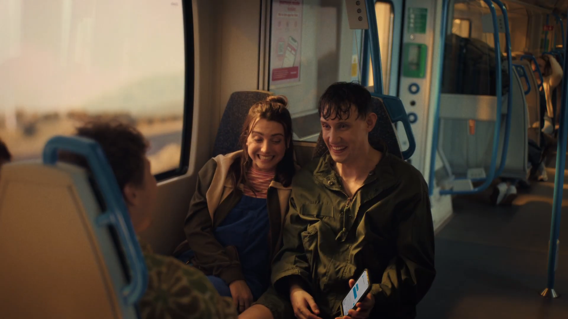 Friends on the Thameslink train home at golden hour — Lily smiling next to Callum (post-paddle wet hair) holding his phone, warm light through the window