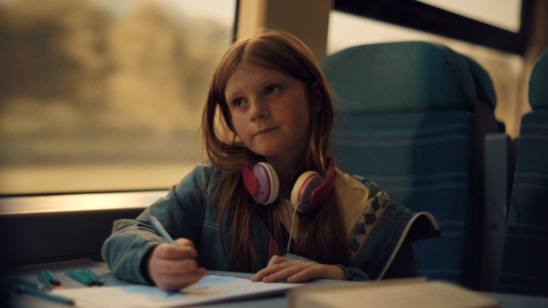 MCU of Sacha at the train table seat drawing in a sketchpad with felt-tip pens — headphones around her neck, golden hour landscape softly out the window