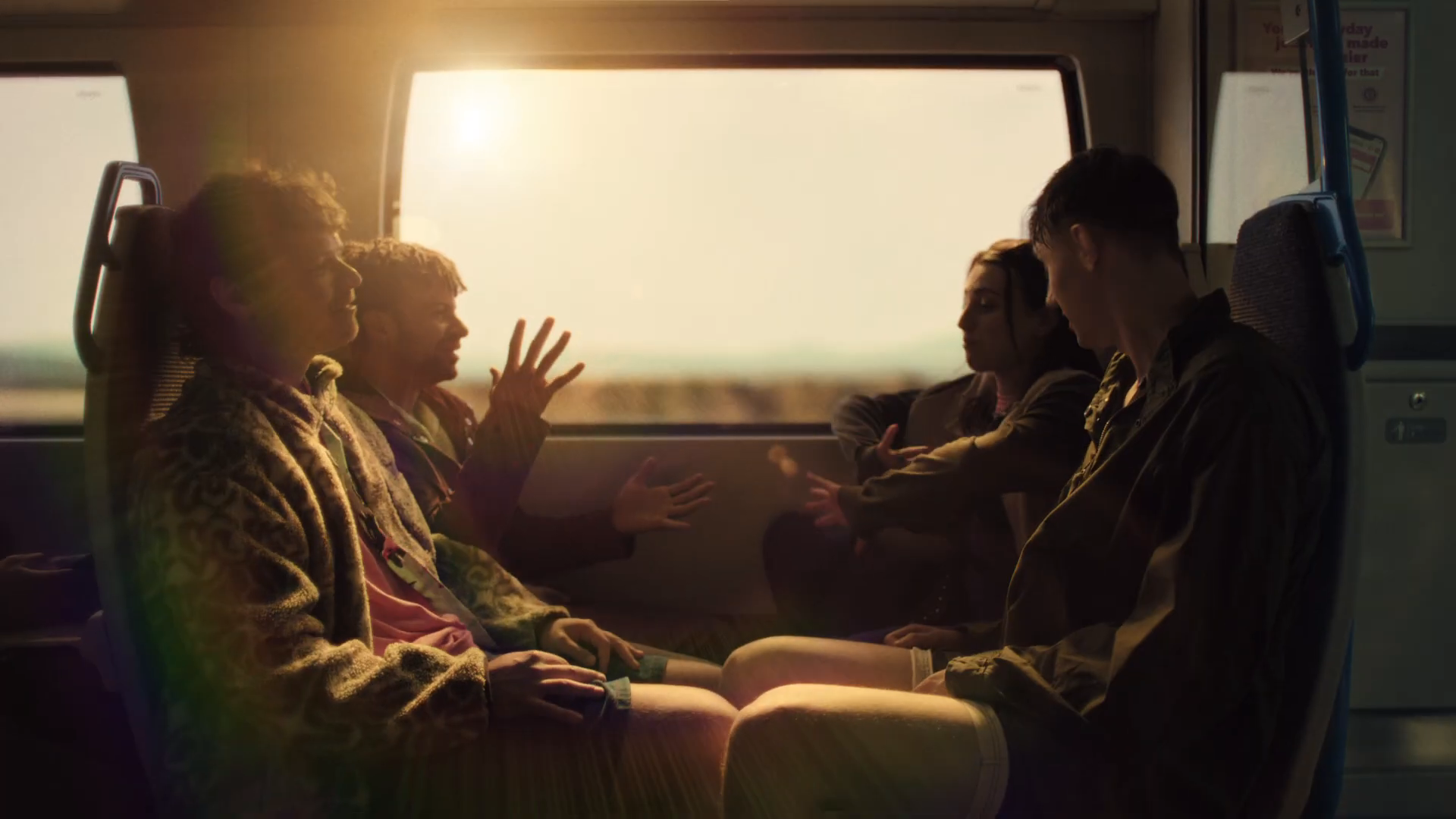 Golden hour ensemble wide on the Thameslink train home — friends animatedly recapping the day with hand gestures mid-storytelling, sun blasting through the panoramic window in lens-flare silhouette