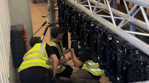 BTS — three crew in hi-vis vests crouching to assemble the LED panel wall row, Thameslink train carriage looming behind, the build-out moment of the volume