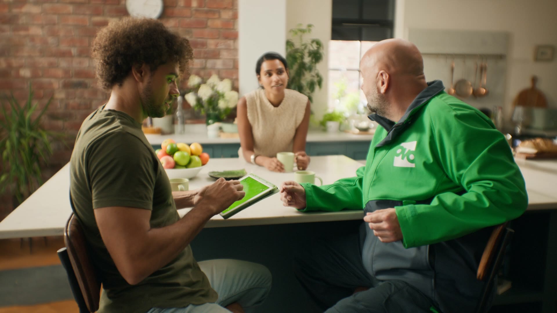 OVO engineer in green branded jacket talking to a couple across a kitchen island, exposed-brick wall behind.