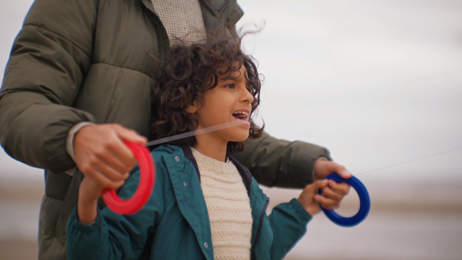 Smiling curly-haired child in teal raincoat, parent leaning in behind in a green puffer.
