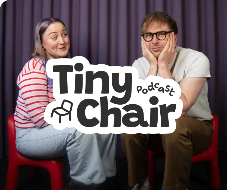 Two people sit on red chairs with a look of amusement. A logo reading Tiny Chair Podcast is centered. The background is a purple curtain
