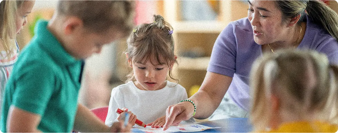 A teacher assists a young girl with a coloring activity at a table, surrounded by other children engaged in similar tasks
