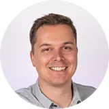 Eine lächelnde Person in einem hellen Hemd vor hellviolettem Hintergrund. Die Stimmung ist freundlich und einladend.