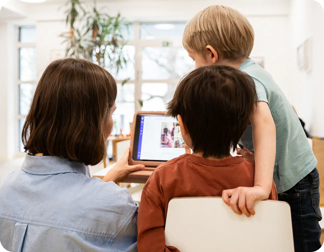 Eine Frau zeigt zwei kleinen Jungen in einem hellen Raum etwas auf einem Tablet. Die Jungen wirken fasziniert, was eine Atmosphäre der Neugier und des Lernens schafft.