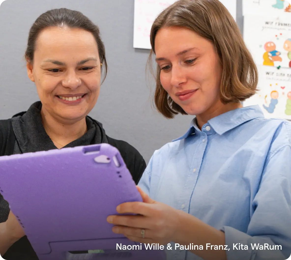 Zwei Frauen lächeln, während sie auf ein Tablet in einer violetten Hülle schauen. Sie stehen vor einer Wand mit bunten Zeichnungen, was eine fröhliche, gemeinschaftliche Atmosphäre vermittelt.