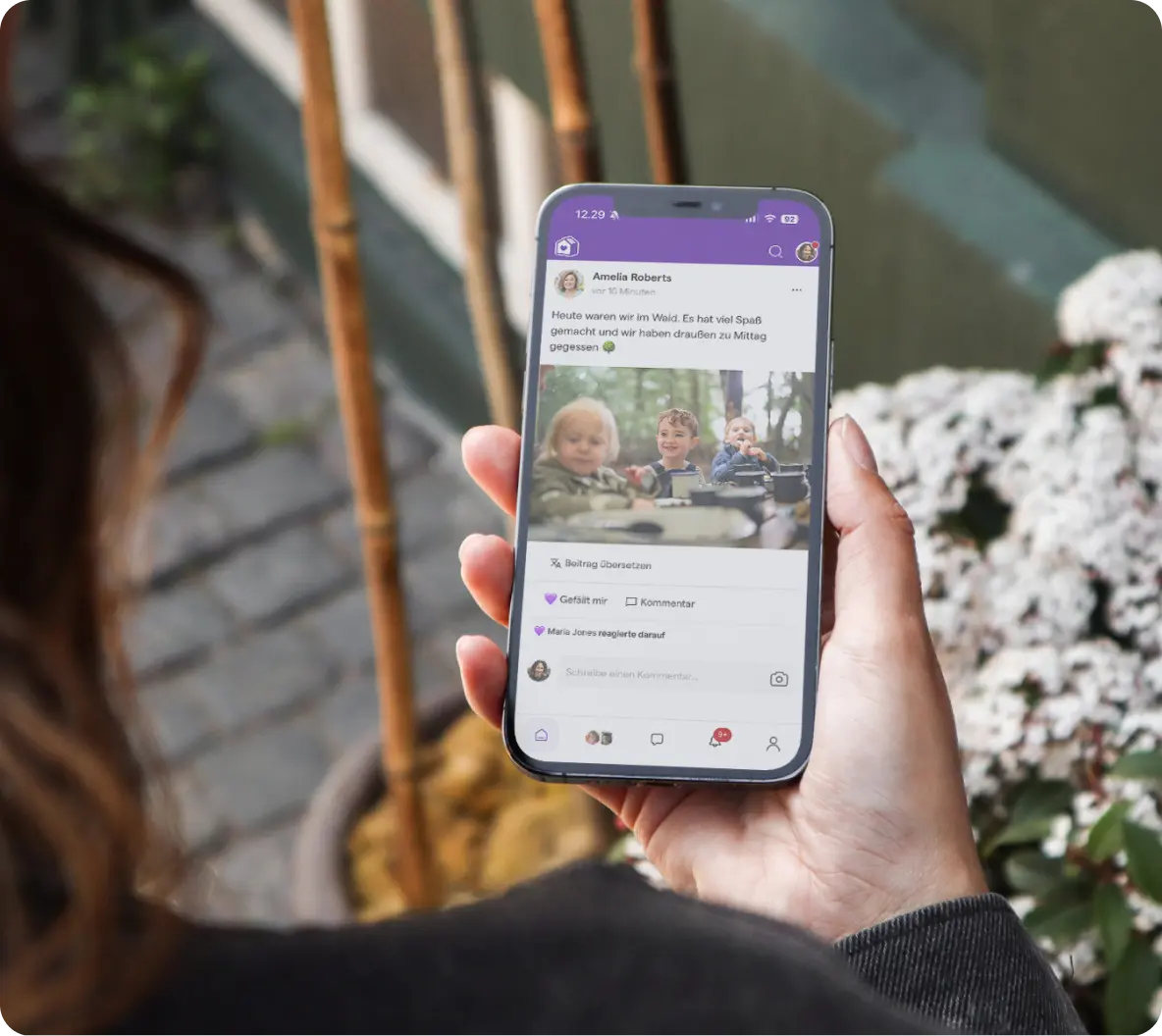 Eine Person hält ein Smartphone in der Hand, auf dem ein Social-Media-Beitrag mit Kindern zu sehen ist, die draußen spielen. Die Szenerie wirkt ungezwungen und fröhlich, umgeben von Blumen.