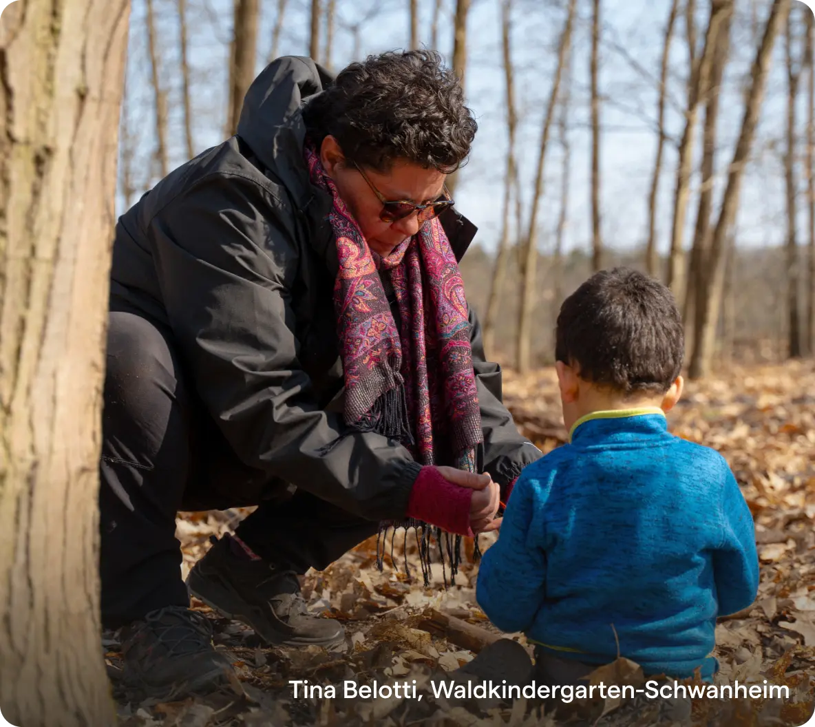 Eine Frau mit buntem Schal und Sonnenbrille kniet im Wald und beschäftigt sich mit einem Kind in einer blauen Jacke. Das Sonnenlicht fällt durch die kahlen Bäume und schafft eine warme, behagliche Atmosphäre.
