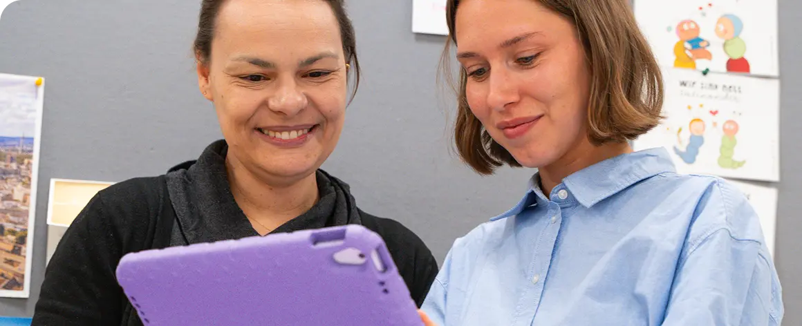 Zwei Frauen schauen gemeinsam auf ein Tablet und lächeln in einer freundlichen Büro- oder Kita-Umgebung. Im Hintergrund hängen Zeichnungen an der Wand.