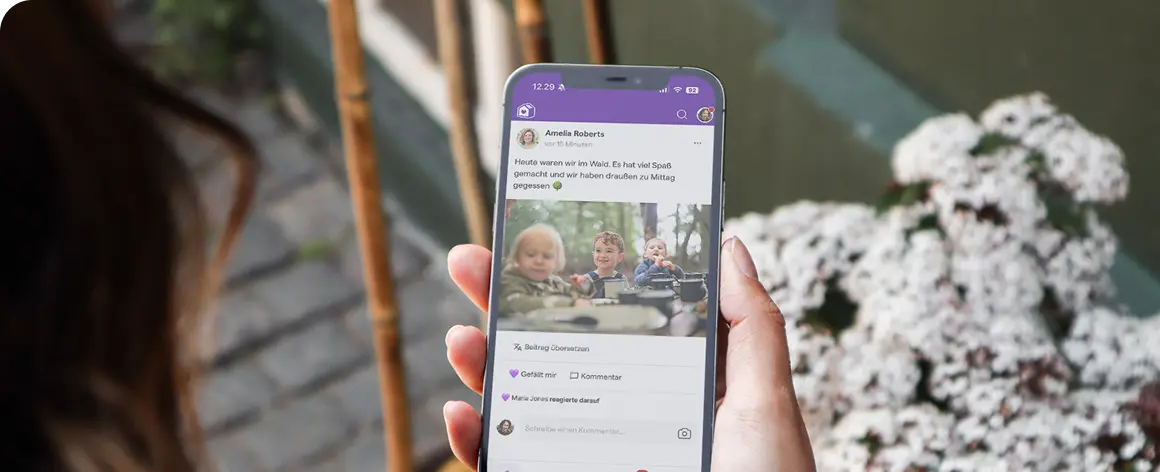 Eine Person hält ein Smartphone in der Hand, auf dem ein Social-Media-Beitrag mit Kindern zu sehen ist, die draußen spielen. Die Szenerie wirkt ungezwungen und fröhlich, umgeben von Blumen.