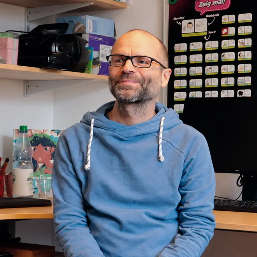 Porträt eines lächelnden Mannes mit Brille und blauem Kapuzenpullover in einem Büro oder Therapieraum. Im Hintergrund sind Regale, ein Computerbildschirm und Kommunikationskarten an einer schwarzen Tafel zu sehen.