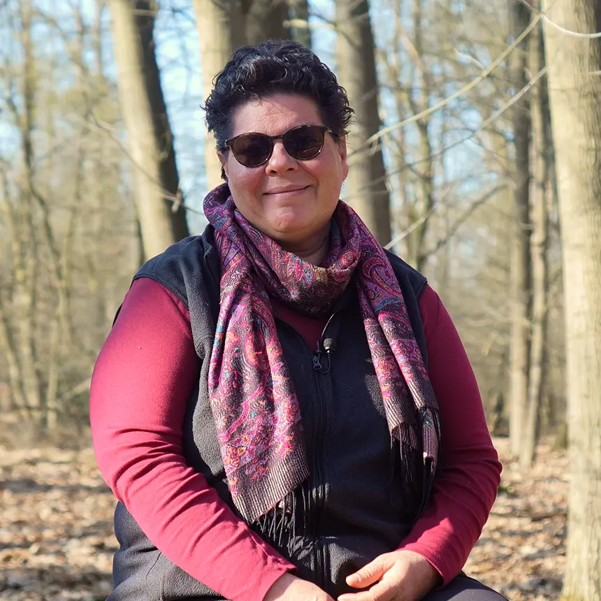 Porträt einer lächelnden Person mit Sonnenbrille und gemustertem Schal in einem sonnigen Wald. Die Person trägt ein bordeauxfarbenes Oberteil und eine schwarze Weste, während kahle Bäume den natürlichen Hintergrund bilden.