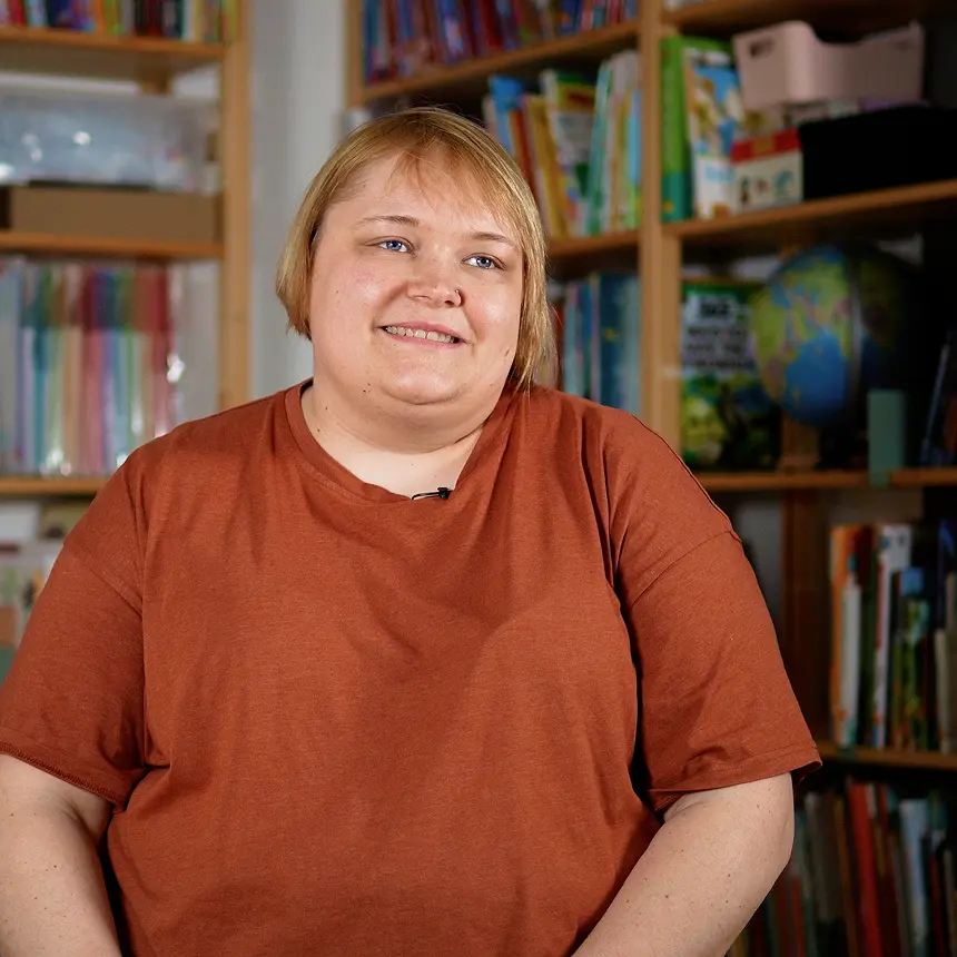 Porträt einer lächelnden Person mit kurzem blondem Haar und braunem T-Shirt vor einem Bücherregal. Im Hintergrund sind Bücher, ein Globus und verschiedene Lernmaterialien in einem wohnlichen Raum zu sehen.