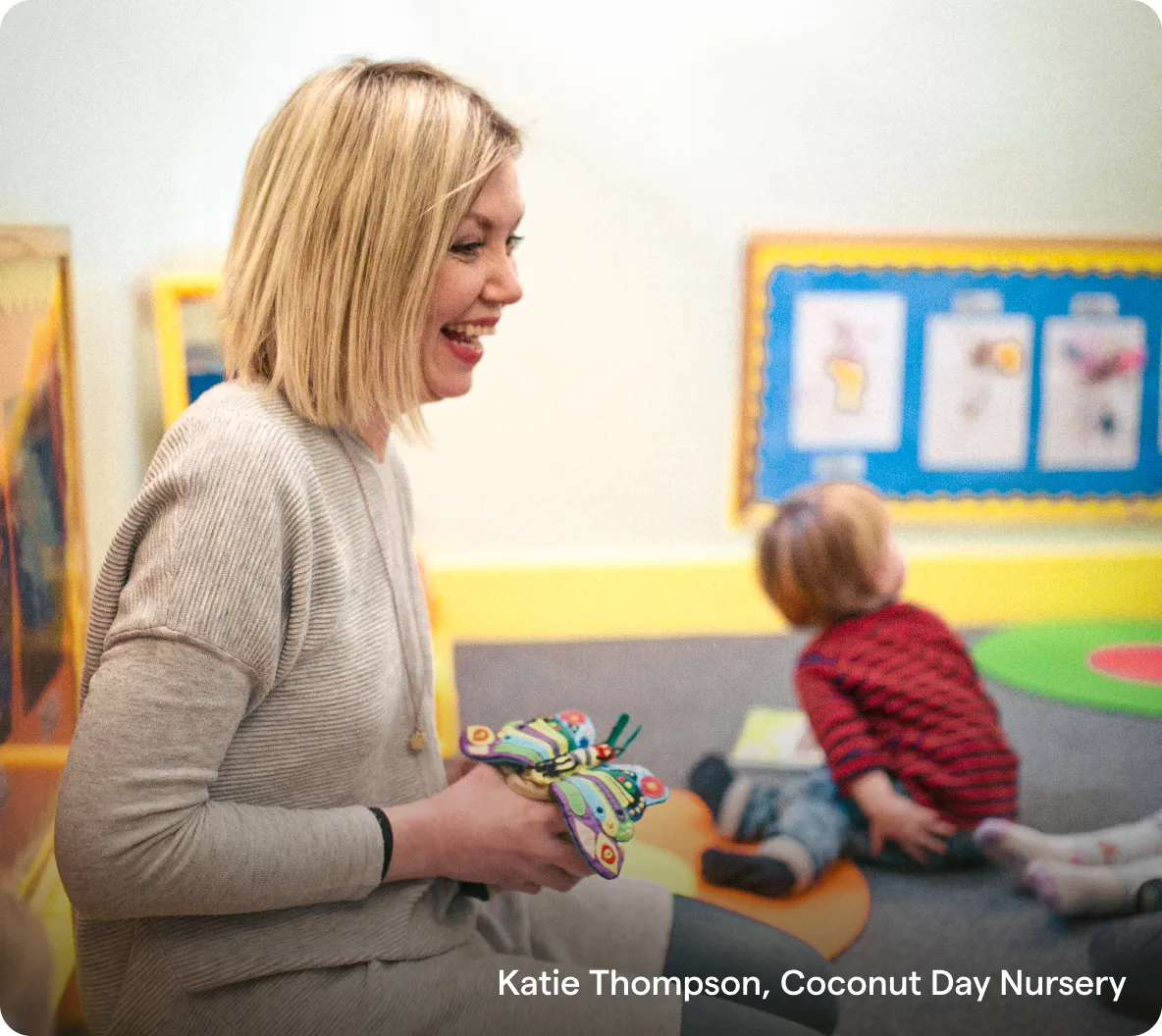 Woman smiling while holding a colourful butterfly toy with children playing in nursery background.