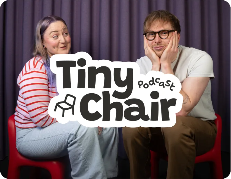 Two Tiny Chair Podcast hosts seated on small red chairs in front of a purple curtain backdrop.