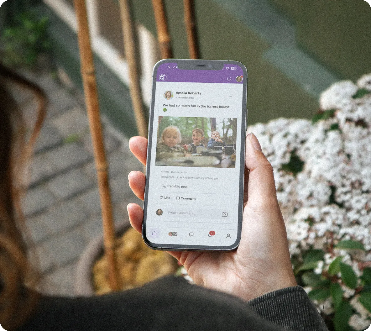 A hand holding a phone with the Famly parent app open, showing a nursery post from educator Amelia Roberts with a photo of children enjoying a forest trip.