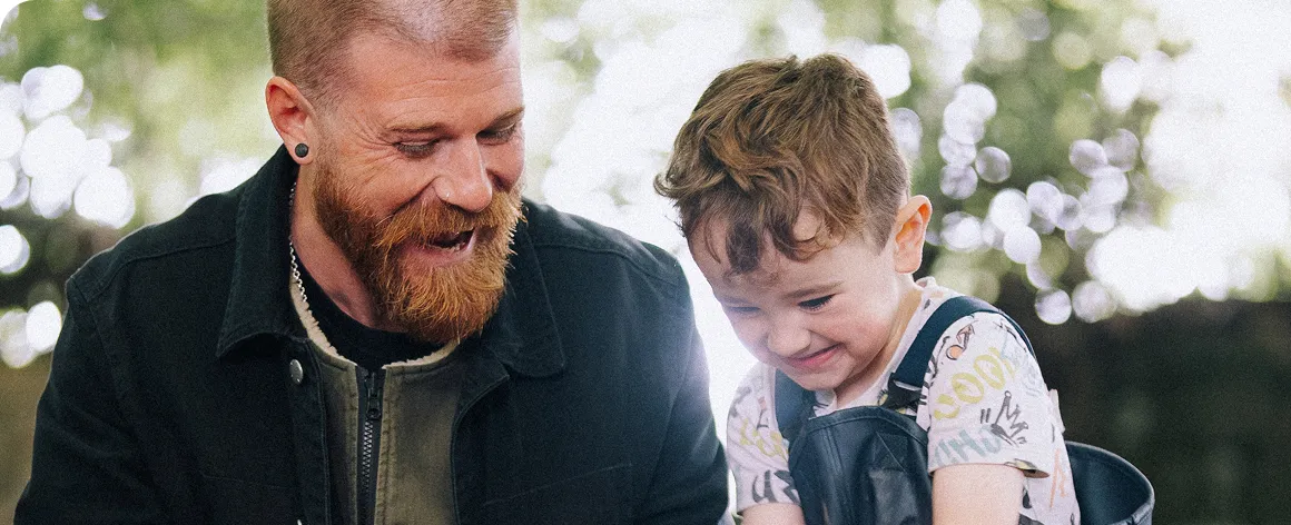Smiling bearded man and young boy looking down together outdoors with blurred lights behind them.