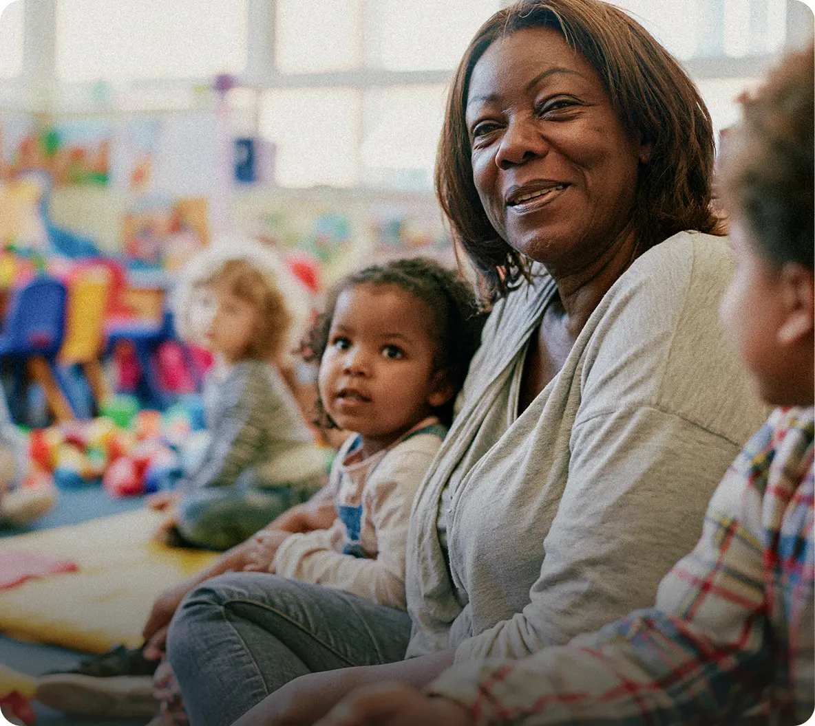 A smiling nursery educator sits with young children during a group activity in a bright, welcoming childcare setting, creating a warm and nurturing learning environment. Children are engaged together, highlighting early years education, care, and social development.