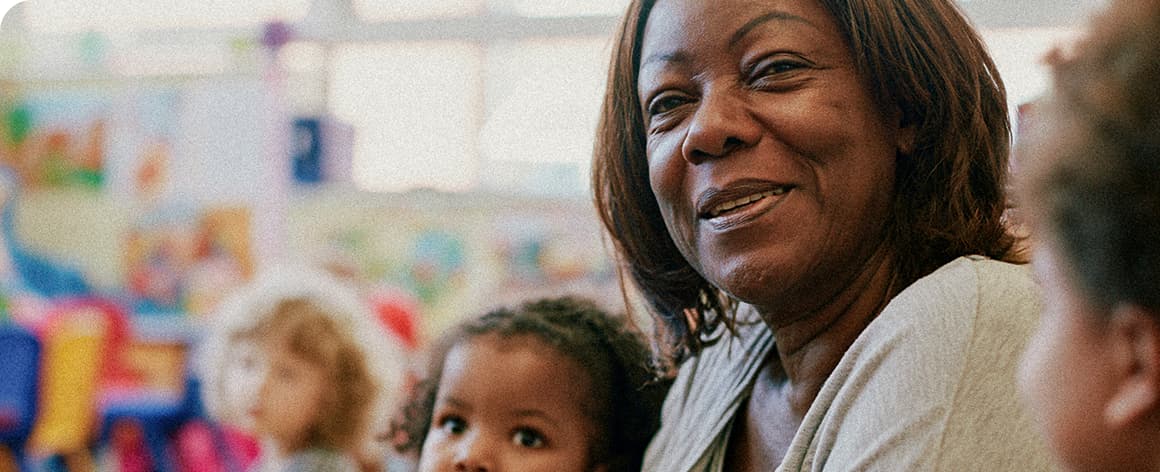 Smiling woman interacting with diverse young children in a colourful classroom setting.