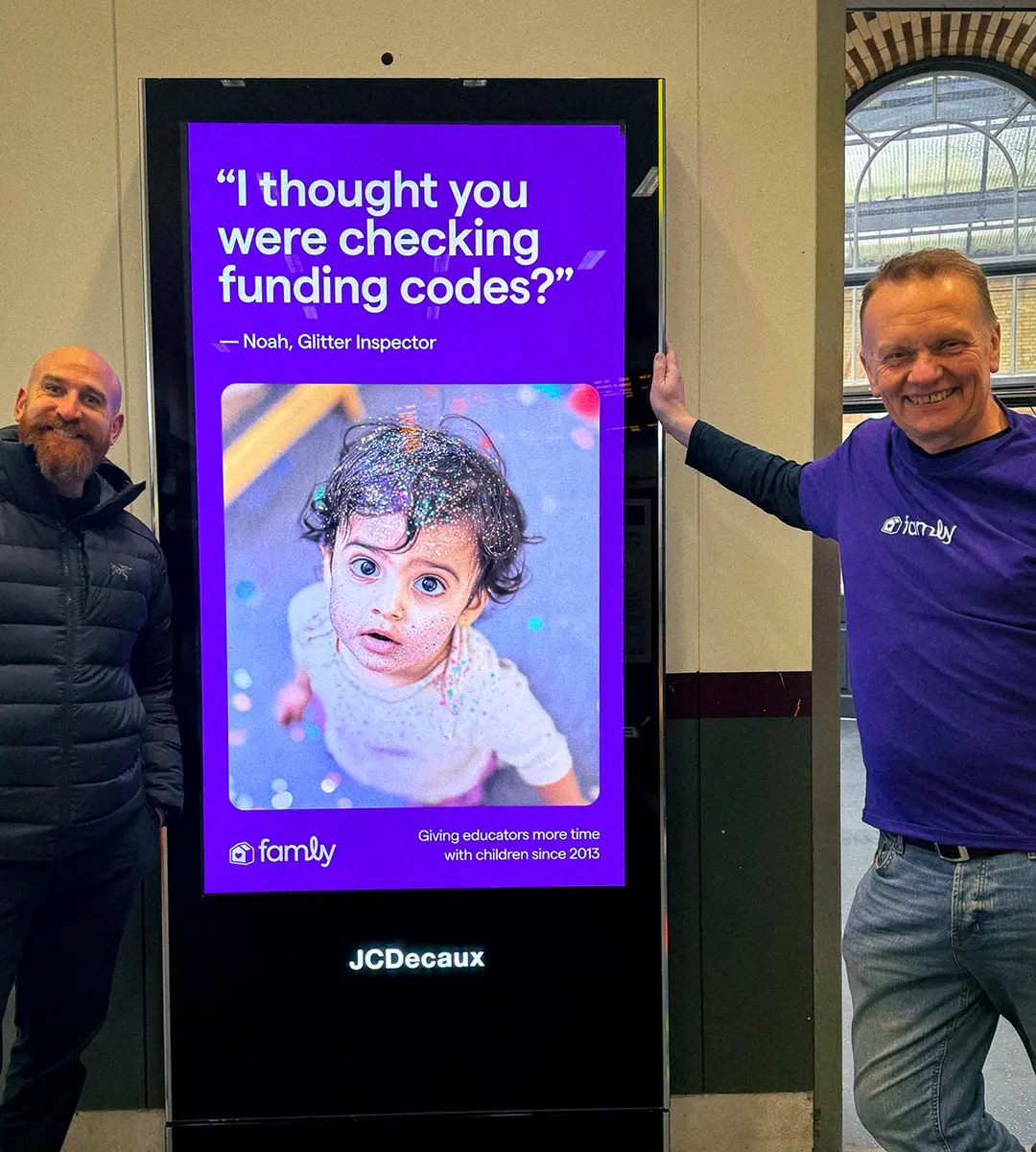 Two Famly colleagues standing either side of a digital billboard showing a Famly advert
