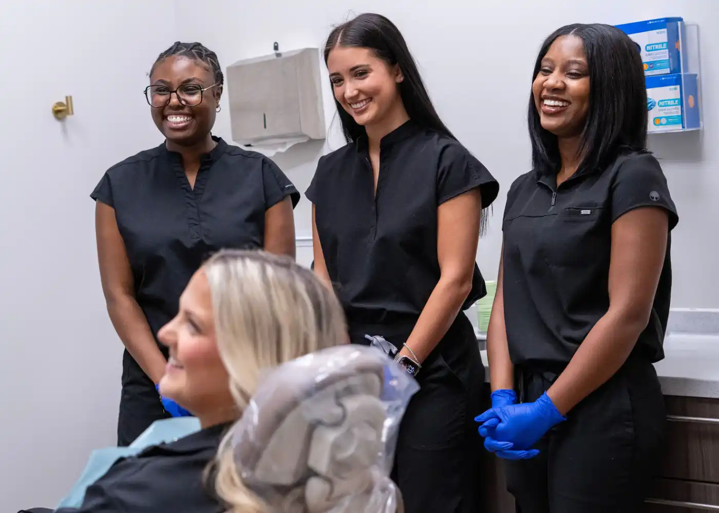Group of five smiling healthcare workers in black scrubs, sitting and standing around a dental chair in a clinic.