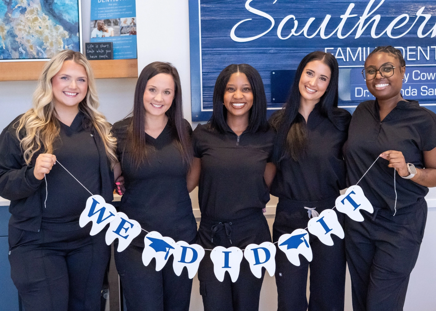 Group of five smiling healthcare workers in black scrubs, sitting and standing around a dental chair in a clinic.