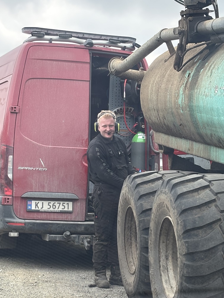 Andreas står på utsiden av arbeidsbilen og reparerer ei gjødselvogn