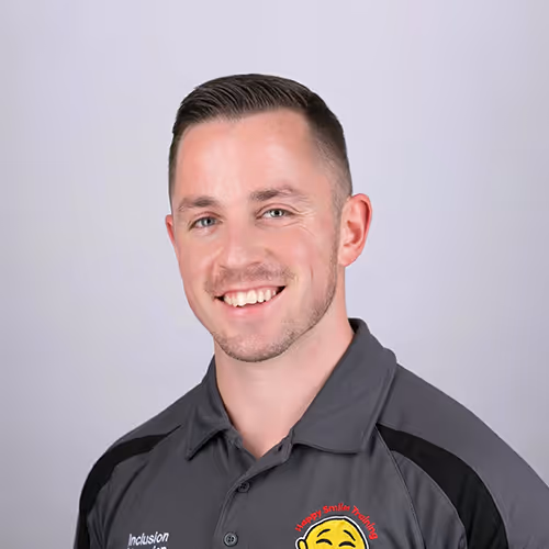 A man with short brown hair is smiling at the camera. He is wearing a grey and black collared shirt with an embroidered logo that features a smiling face and the words “Inclusion” and “Happy Smiles Training.” The background is a plain light grey.