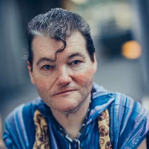 A man with pale skin, dark wavy hair, and a distinctive curl on his forehead is looking directly at the camera with a slight smile. He is wearing a colourful striped shirt with patterned details. The background is softly blurred, suggesting an urban setting.