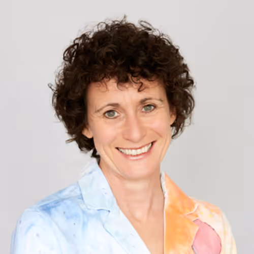 A woman with short curly brown hair smiling warmly. She is wearing a colourful shirt with a watercolour-style pattern in blue, orange, and pink tones. The background is a plain light grey.