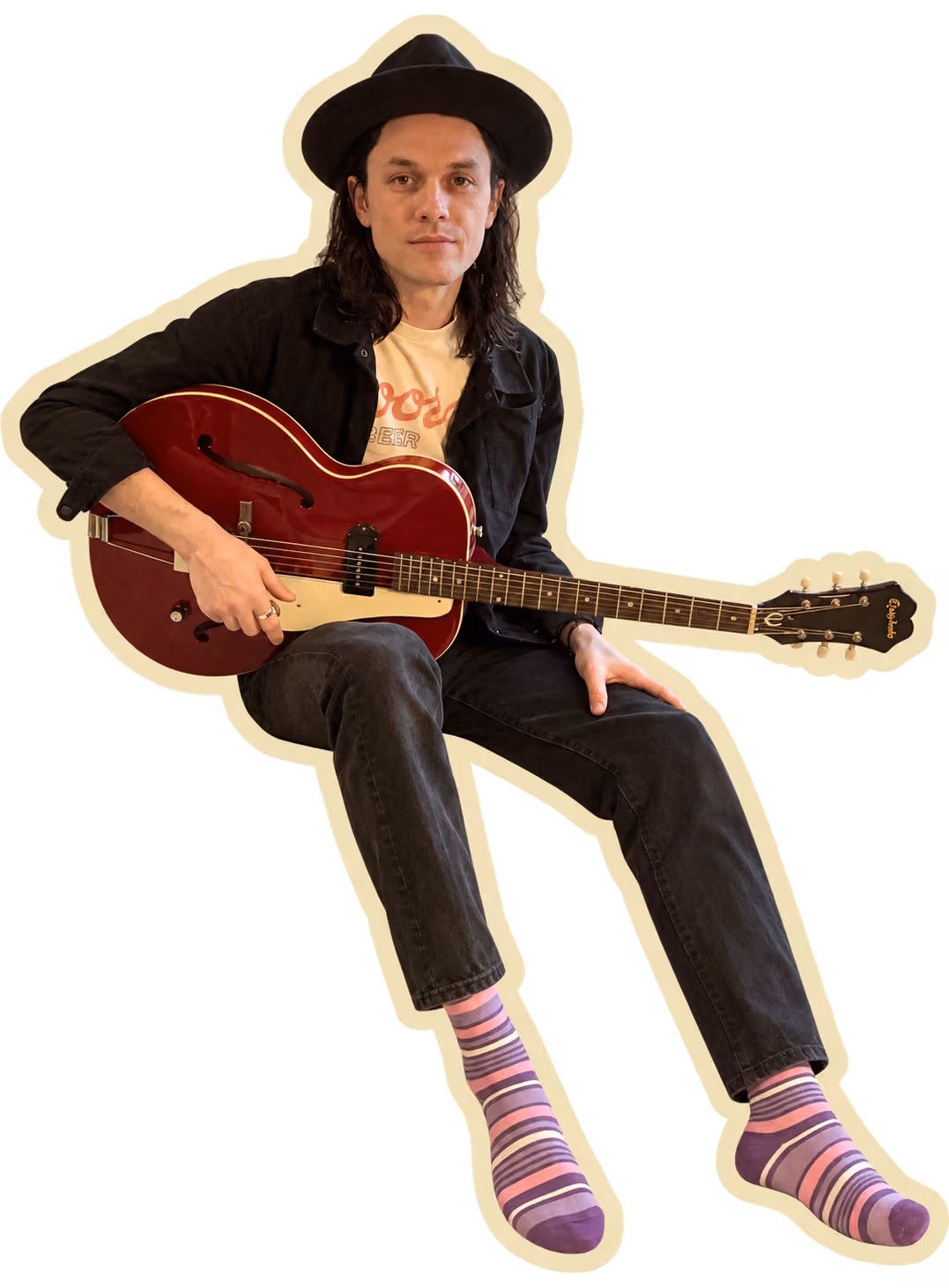 James May seated on a chair holding a red hollow-body guitar, wearing a black wide-brimmed hat and dark clothing with purple striped socks.